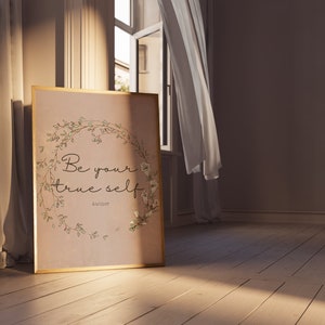 May include: A framed print with a floral wreath and the text "Be your true self" with the author "Aulthor" below. The print is on a light brown background and has a gold frame. The print is sitting on a wooden floor.