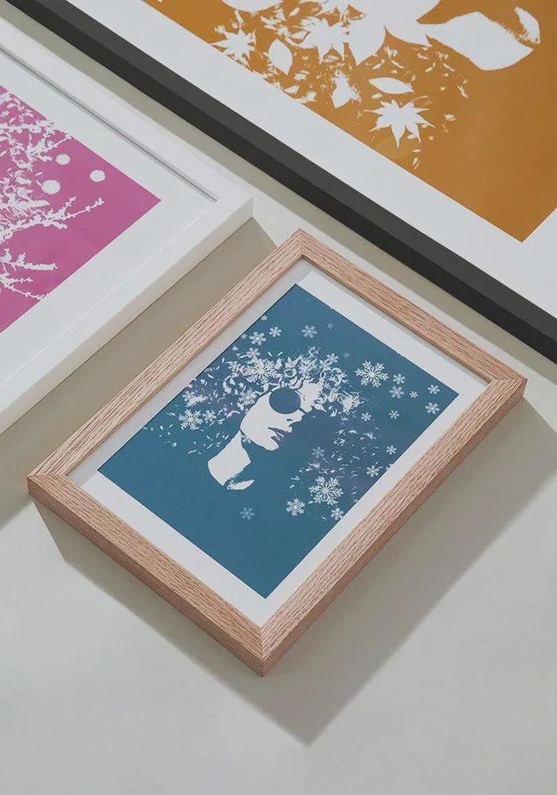Puede incluir: Impresiones art&iacute;sticas enmarcadas con tem&aacute;tica invernal. Una impresi&oacute;n tiene un fondo azul con la silueta blanca de una persona con gafas de sol y copos de nieve. Otras impresiones tienen fondos rosas y dorados con dise&ntilde;os florales. Los marcos son de madera clara.
