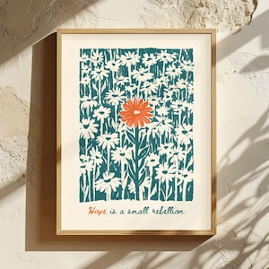 May include: A framed wall art print featuring a field of white flowers against a dark teal background, with one prominent orange flower in the center. The text "Hope is a small rebellion" is printed in orange and dark teal at the bottom.