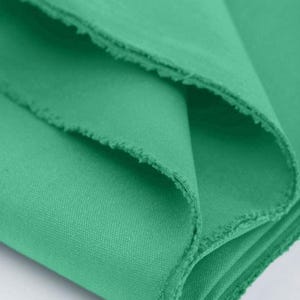 Könnte beinhalten: Ein gefaltetes Stück mintgrüner Stoff mit einer sichtbaren Webstruktur und ausgefransten Kanten entlang der Falten. Das Material ist blickdicht und weist eine gleichmäßige, leuchtend grüne Farbe auf der gesamten Textiloberfläche auf.