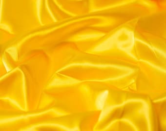 May include: A vibrant yellow fabric with a smooth, lustrous finish. The material is draped in soft, flowing folds that create deep shadows and bright highlights across the surface, emphasizing the reflective quality of the textile texture.