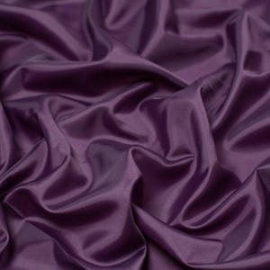 May include: A solid deep purple fabric with a smooth, slightly reflective surface. The material is draped in soft, flowing folds, creating a series of highlights and shadows that emphasize the texture and consistent dark violet color throughout.