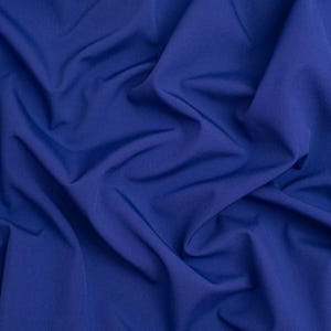 May include: A solid royal blue fabric with a smooth, matte texture. The material is draped in soft, flowing folds that create deep shadows and highlights across the surface, emphasizing the consistent, vibrant blue color throughout the textile.