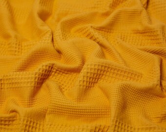 May include: A vibrant mustard yellow fabric featuring a distinct waffle weave texture. The material is draped in soft, flowing folds, highlighting the raised grid pattern and the consistent, deep golden hue across the entire surface of the textile.
