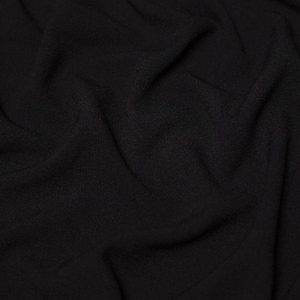 May include: A solid black fabric with a soft, matte texture and a fine, granular surface appearance. The material is draped in loose, flowing folds, creating deep shadows and highlights that emphasise the consistent, dark, and opaque colour.