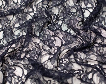 May include: A piece of black lace fabric featuring a detailed floral pattern with intricate petals and leaves. The lace has a fine mesh background and is draped to show the texture and the contrast between the dark thread and the light surface.