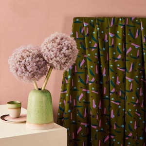 May include: A light green ceramic vase holds two large, spherical, light purple allium flowers. Beside the vase sits a small, matching light green cup on a saucer. A draped fabric features a repeating pattern of dark green, pink, and blue shapes.
