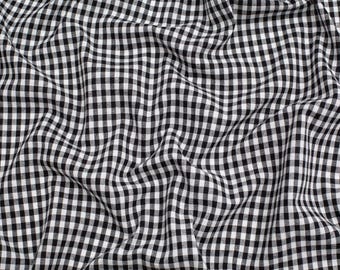 May include: A fabric featuring a classic black and white gingham check pattern. The material is arranged in soft, flowing folds, creating a series of deep shadows and highlights that emphasize the repeating square grid design across the surface.