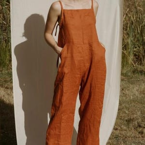 Peut inclure: Une combinaison en lin de couleur rouille avec des bretelles fines et un décolleté carré. La combinaison a deux poches sur le devant et des jambes larges. Le mannequin porte des sandales noires.