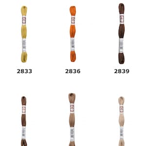 May include: Six skeins of D.M.C. Soft Cotton Embroidery Thread arranged in two rows. The colors include mustard yellow, orange, dark brown, medium brown, light brown, and beige. Each skein features a paper label with the D.M.C. logo and number.