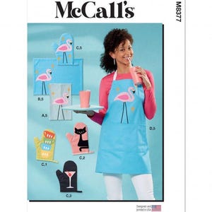 May include: McCall's sewing pattern M8377 featuring kitchen accessories including a light blue apron with a pink flamingo graphic, oven mitts with flamingo, cat, and cocktail designs, and a tea towel with a flamingo print on a light blue background.