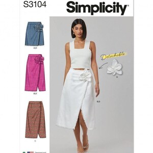 Könnte beinhalten: Simplicity Schnittmuster S3104 zeigt einen Wickelrock mit abnehmbarer Blumenapplikation. Das Muster umfasst Varianten in Jeansblau, leuchtendem Pink und strukturiertem braunem Stoff, dargestellt mit weißem Top und weißem Rock.