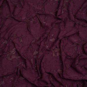 May include: A deep plum purple fabric featuring a textured floral lace pattern. The material is gathered and folded, creating a series of soft, irregular ripples and shadows across the surface that highlight the intricate, raised botanical design.