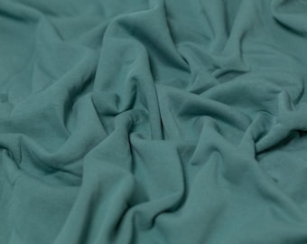 May include: A solid teal fabric with a matte finish, featuring deep folds and soft drapes that create a textured, layered appearance across the surface. The material has a consistent, muted blue-green color throughout the entire visible area.