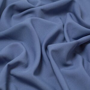 Puede incluir: Una tela de color azul bígaro sólido con una textura suave y mate. El material está drapeado en pliegues suaves y fluidos, creando sombras y reflejos profundos en la superficie que enfatizan el color consistente y el tejido uniforme.