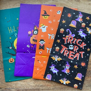 Könnte beinhalten: Vier Halloween-Papiertüten mit verschiedenen Designs. Die erste Tüte ist grün mit weißen Sternen und dem Text "HALLOWEEN PARTY". Die zweite Tüte ist lila mit einer Comic-Katze und dem Text "HAPPY HALLOWEEN". Die dritte Tüte ist orange mit einem Comic-Geist und dem Text "TRICK OR TREAT". Die vierte Tüte ist schwarz mit weißen Sternen und dem Text "TRICK OR TREAT".