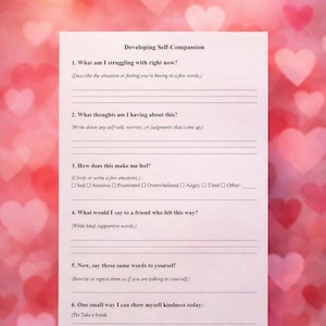 May include: A white paper titled "Developing Self-Compassion" with prompts for self-reflection. The prompts include questions about current struggles, thoughts, feelings, and self-kindness. The background has a blurred pink and red heart pattern.