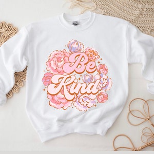 Puede incluir: Sudadera blanca con un diseño floral rosa y naranja y el texto "Be Kind" en una fuente retro.