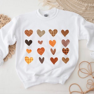 Könnte beinhalten: Weißer Pullover mit einem Muster aus braunen, orangen und gelben Herzen. Die Herzen sind in verschiedenen Formen und Größen und haben verschiedene Muster, darunter Punkte, Karos und Streifen.