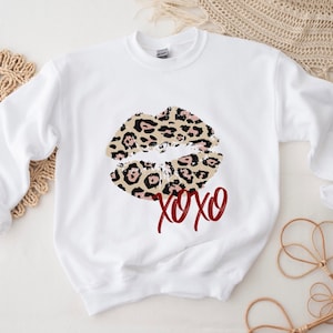 Puede incluir: Sudadera blanca con un diseño de labios de estampado de leopardo y el texto "XOXO" en rojo.