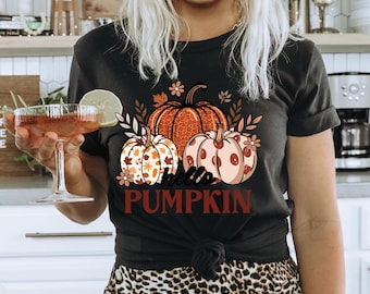 May include: A black t-shirt with a graphic of three pumpkins and the text "hello pumpkin". The pumpkins are orange, brown, and white with fall-themed patterns. The model is wearing a brown hat and leopard print pants.