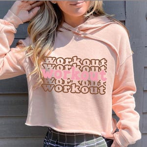 Könnte beinhalten: Ein rosa, kurzer Hoodie mit einem viermal wiederholten "Workout"-Motiv in Braun und Rosa.