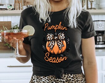May include: A black t-shirt with a pumpkin graphic and the text "Pumpkin Season". The pumpkin has a leopard print bow. The person wearing the shirt is also wearing a brown hat and leopard print pants.