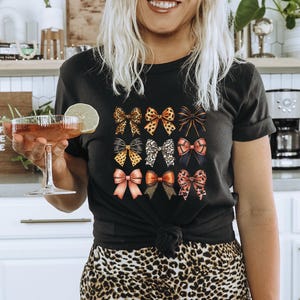 Könnte beinhalten: Eine Frau trägt ein schwarzes T-Shirt mit einem bunten Schleifen-Druck und eine Hose mit Leopardenmuster. Sie hält ein Glas Rotwein in der Hand und trägt einen braunen Hut.