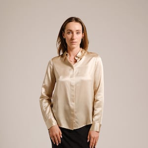 Puede incluir: Una mujer con una camisa de satén dorada con cuello abotonado y mangas largas. También lleva una falda negra.