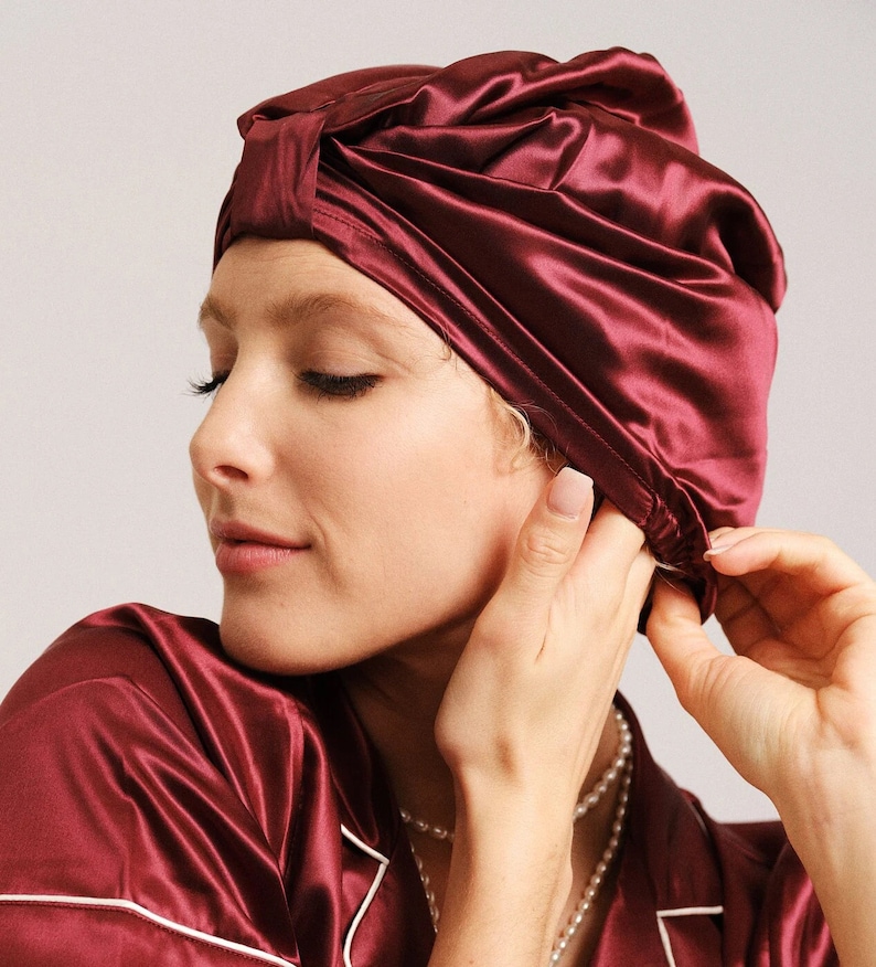 A burgundy silk sleep cap with a gathered design. The cap is worn by a person with blonde hair and a pearl necklace.