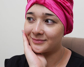 Op de afbeelding: Een vrouw draagt een roze satijnen slaapmuts. De muts is bovenop haar hoofd in een knoop geknoopt. Ze draagt een zwart shirt en een zilveren ketting met een blauwe edelsteen hanger.