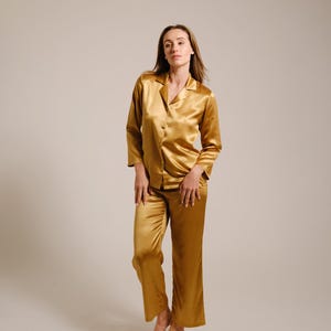 May include: A golden-yellow silk pajama set. The long-sleeved button-down shirt has a notched collar. The matching pants are loose-fitting. The fabric has a subtle sheen. The model is standing barefoot.
