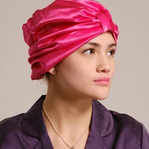 Pu&ograve; includere: Una donna indossa un turbante di raso rosa brillante con una collana d'argento con un ciondolo a forma di uccello.