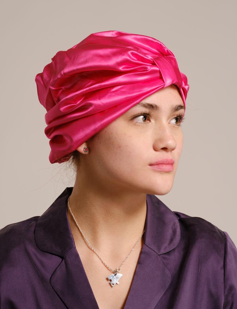 Pu&ograve; includere: Una donna indossa un turbante di raso rosa brillante con una collana d'argento con un ciondolo a forma di uccello.