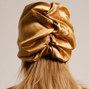 Pu&ograve; includere: Un copricapo da notte in raso dorato con un design raccolto e gonfio. Il copricapo &egrave; indossato da una persona con capelli lunghi e castano chiaro.
