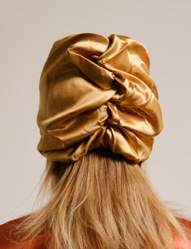 Pu&ograve; includere: Un copricapo da notte in raso dorato con un design raccolto e gonfio. Il copricapo &egrave; indossato da una persona con capelli lunghi e castano chiaro.