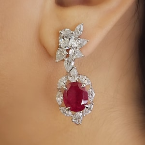 Könnte beinhalten: Ein Paar Hängeohrringe mit einem großen roten Rubin-Edelstein, der von kleineren klaren Edelsteinen umgeben ist. Die Ohrringe sind aus Silber und haben ein florales Design.