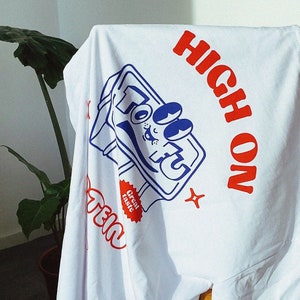 May include: White t-shirt featuring a blue and white cartoon illustration of a can of soda. The text "HIGH ON" is printed in red above the can and "TEI" is printed in red below the can. A red starburst sticker with the text "Great taste" is on the can.