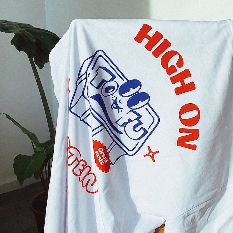 May include: White t-shirt featuring a blue and white cartoon illustration of a can of soda. The text "HIGH ON" is printed in red above the can and "TEI" is printed in red below the can. A red starburst sticker with the text "Great taste" is on the can.