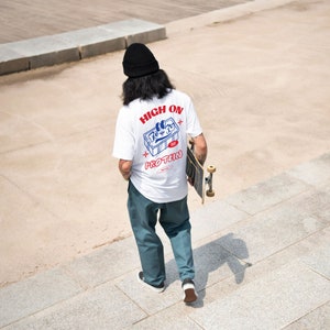 May include: A person wearing a white t-shirt with a blue and red graphic that says "High on Protein" and a blue and white illustration of a protein bar. The person is holding a skateboard.