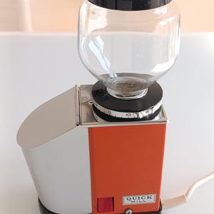 Può includere: Un macinacaffè Quick Mill arancione e argento con una tramoggia per chicchi in vetro trasparente e un coperchio nero. Il macinacaffè ha un pulsante di accensione rosso e un quadrante per le impostazioni di macinatura. Il cavo è bianco.