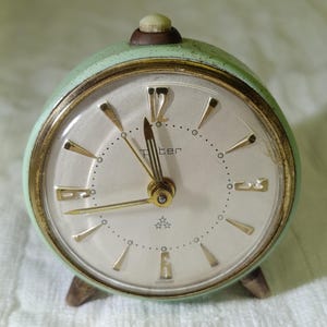 Könnte beinhalten: Ein Vintage-Wecker in Mintgrün mit goldfarbener Lünette und Zeigern. Das Zifferblatt ist weiß mit schwarzen Ziffern und dem Markennamen "Piter". Die Uhr hat einen kleinen, runden Knopf oben und drei kleine Füße.