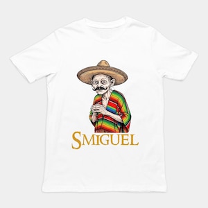 Peut inclure: Un t-shirt blanc à manches courtes avec une illustration graphique représentant une créature pâle et fine portant un sombrero marron, une moustache noire et un poncho rayé multicolore. Le texte SMIGUEL est imprimé en doré en dessous.