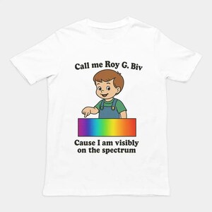 Op de afbeelding: Een wit t-shirt met een cartoonillustratie van een kind in een groen shirt en blauwe tuinbroek dat naar een horizontale regenboogrechthoek wijst. De tekst luidt Call me Roy G. Biv erboven en Cause I am visibly on the spectrum eronder.