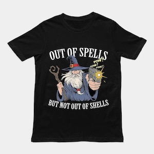 Op de afbeelding: Een zwart t-shirt met een afbeelding van een tovenaar in een blauwe mantel en hoed die een houten staf in de ene hand en een pistool in de andere hand houdt. De tekst luidt OUT OF SPELLS BUT NOT OUT OF SHELLS met POW! POW! POW!.