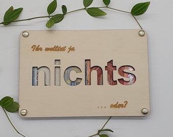 May include: A light beige wooden sign with the text "Ihr wolltet ja nichts... oder?" in a light brown colour. The word "Nichts" is cut out and a euro bill is visible through the cutout.