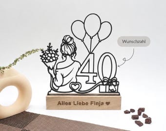 May include: A black wooden 40th birthday cake topper. The topper features a silhouette of a woman holding flowers and balloons. The base is engraved with the text "Alles Liebe Finja".