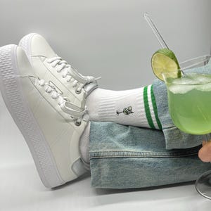 Könnte beinhalten: Weiße Turnschuhe mit weißen Schnürsenkeln und dicker Sohle. Weiße Socken mit grünen Streifen und einer Cocktailgrafik. Ein grüner Cocktail mit Limettenspalten-Garnitur in einem durchsichtigen Glas.