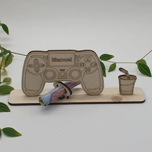 Könnte beinhalten: Ein heller Holzständer mit einer lasergeschnittenen Spielcontroller-Form, in deren Mitte der Name Manuel eingraviert ist. Der Ständer enthält eine kreisförmige Halterung für einen gerollten Geldschein und einen kleinen Ausschnitt eines Bechers.