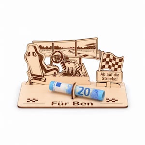 May include: A laser-cut wooden display featuring a racing simulator setup with a seat, steering wheel, and monitors. A checkered flag sign reads Ab auf die Strecke! A rolled 20 Euro bill sits in a holder. The base is engraved with Für Ben.
